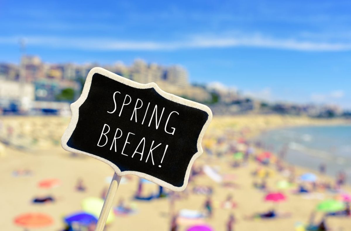 closeup of a black signboard with the text spring break written in it, in front of a blurred beach with many people bathing and sunbathing