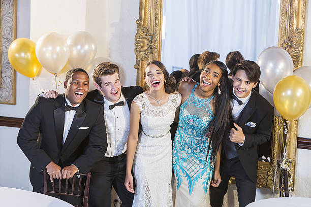 A group of five teenagers dressed in formalwear  They are at a special event, a party or prom, standing together in a row, laughing and smiling.